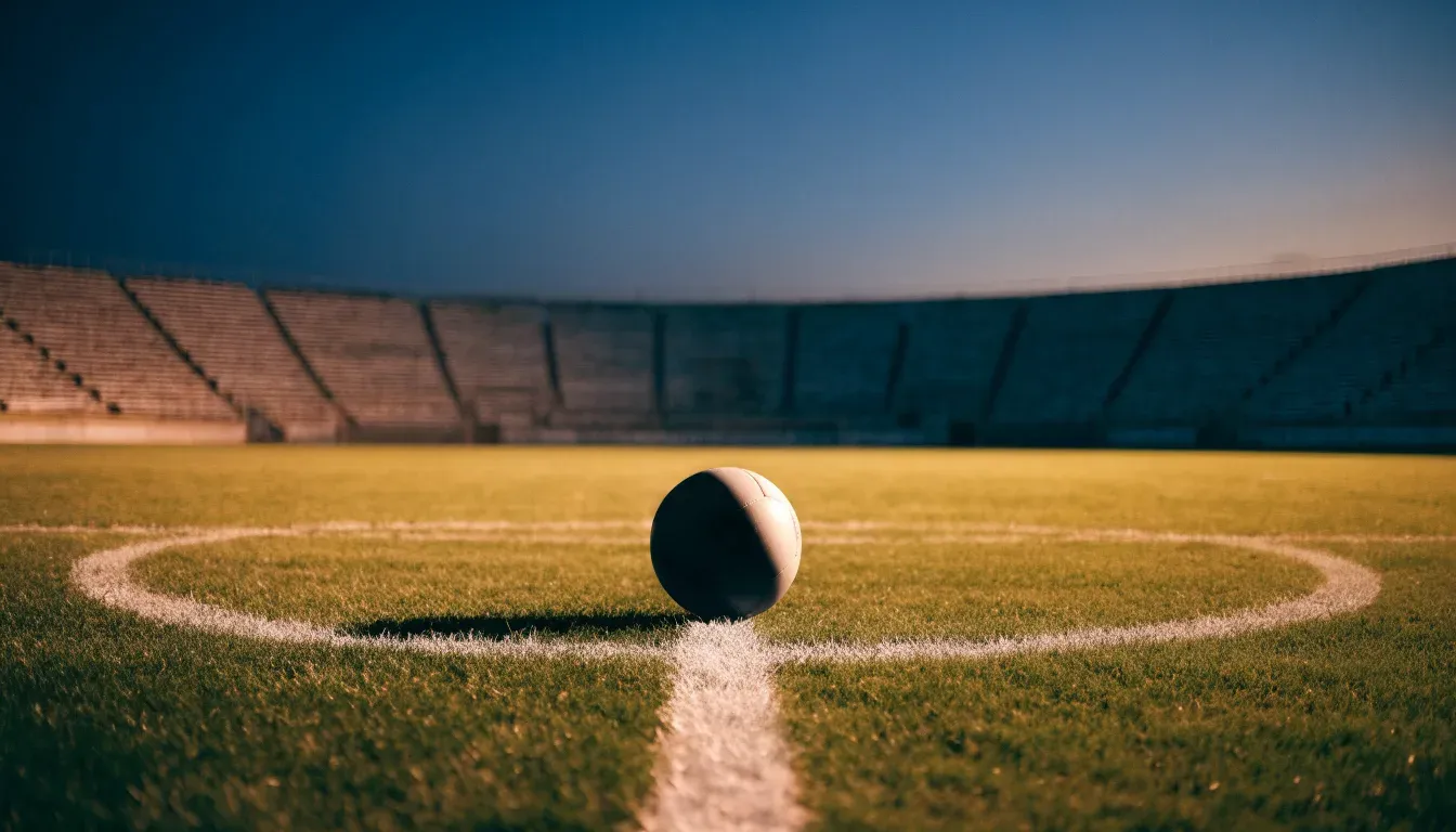 Ein Fussball liegt auf dem Mittelkreis eines leeren Rasenplatzes, dahinter ragen leere Tribünenreihen in den Abendhimmel.