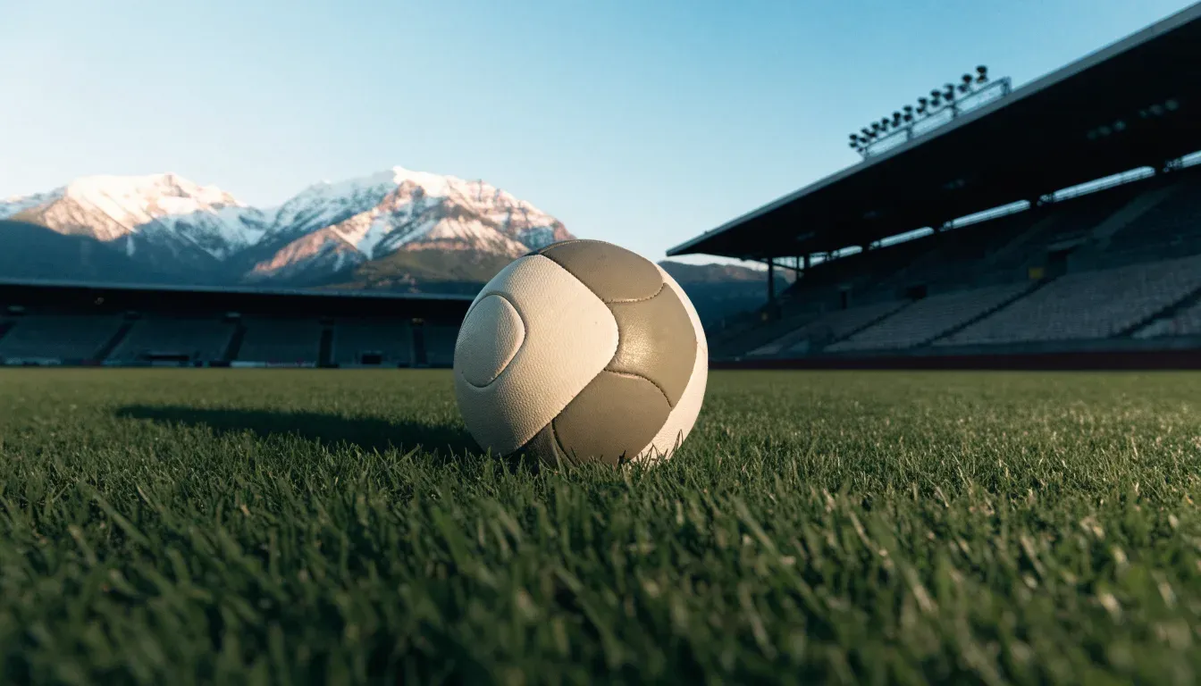 Ein Fussball liegt auf dem Rasen eines leeren Stadions am frühen Abend, dahinter erheben sich schneebedeckte Berge unter klarem Himmel.