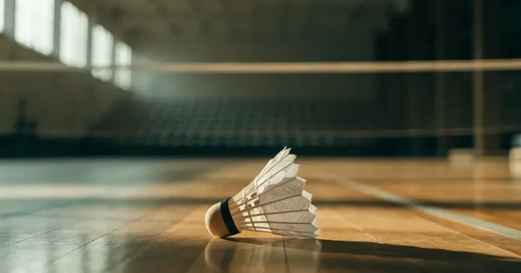 Ein Federball liegt auf dem Boden einer leeren Sporthalle, das Netz verschwimmt im Hintergrund, Licht fällt durch hohe Fenster.