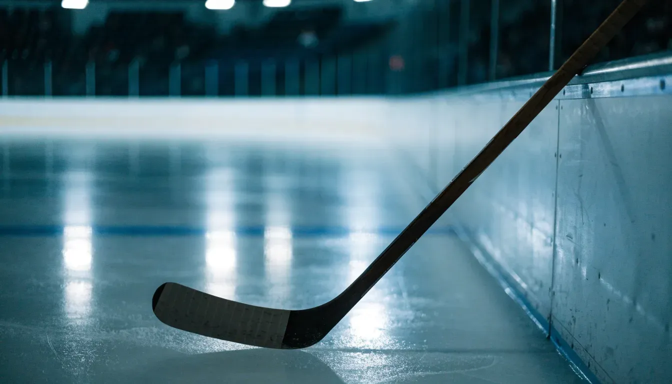 Ein Eishockeyschläger lehnt an der Bande, in der Ferne liegt die hell beleuchtete Eisfläche einer Halle unter Flutlicht.