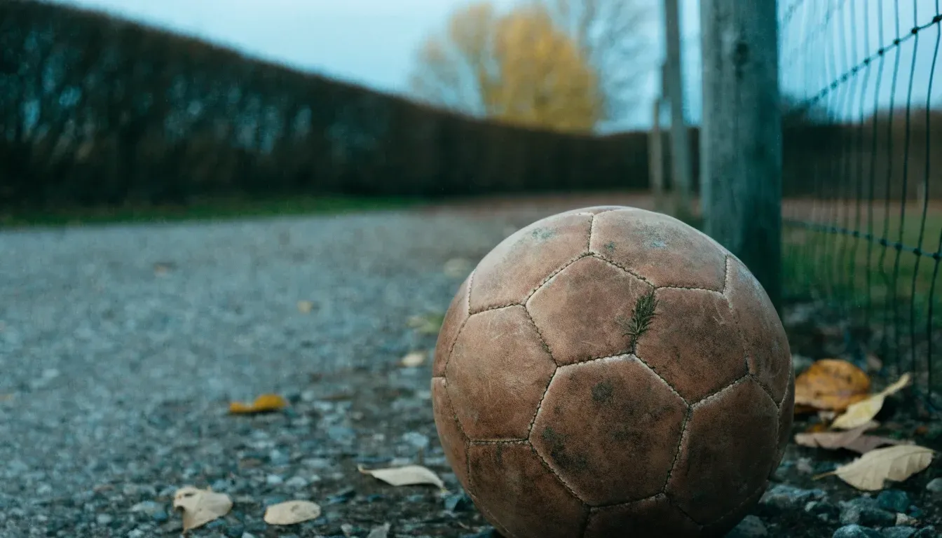 Ein abgenutzter Fussball liegt auf einem Kiesweg neben dem Eingang eines Vereinsplatzes, Laub liegt auf dem Boden.