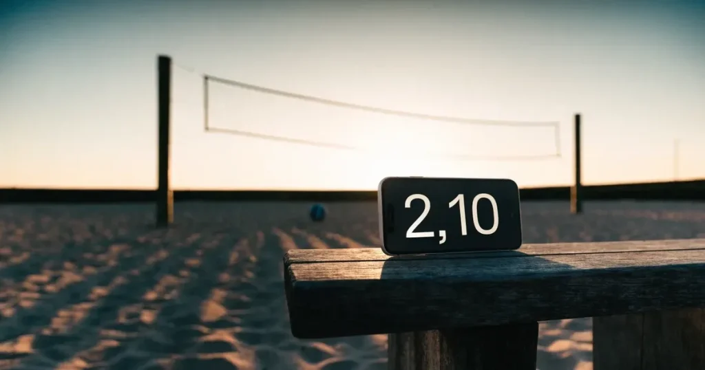 Ein Volleyball liegt im Sand eines Beachplatzes, zwischen zwei Pfosten ist ein Netz gespannt, tiefe Abendsonne wirft lange Schatten.