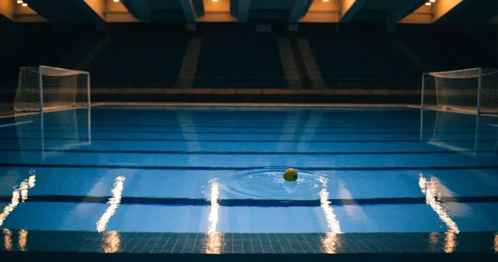 Ein Wasserballbecken am Abend mit zwei schwimmenden Torrahmen an den Enden, ruhiges blaues Wasser, leere Ränge.