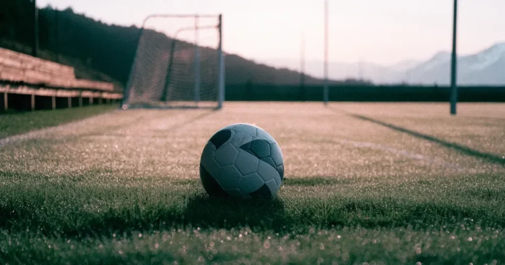 Ein Fussball liegt am frühen Morgen auf dem Rasen eines leeren Fussballfeldes, dahinter eine kleine Tribüne und schneebedeckte Berge.