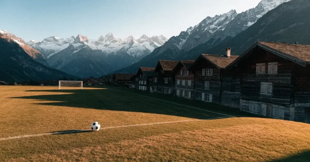 Ein Dorf-Sportplatz mit einem Ball am Strafraumrand, dahinter schneebedeckte Alpengipfel im klaren Morgenlicht