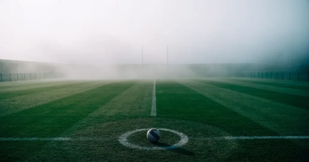 Ein Fussball liegt auf dem Anstosspunkt eines leeren Rasenplatzes im dichten Morgennebel.